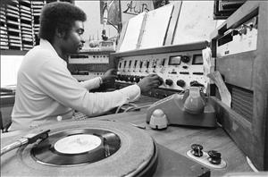 A Black man with sideburns and a full head of hair at 70s era radio studio controls. To his right is a rotary dial telephone and a turntable. 